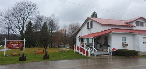 A white building with a red roof, labeled "Wager's Cider Mill," surrounded by trees and a grassy area on a cloudy day.