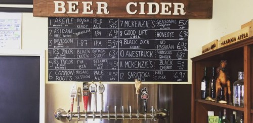 A chalkboard menu displaying various beers and ciders with prices, alongside a bar with taps and bottles.