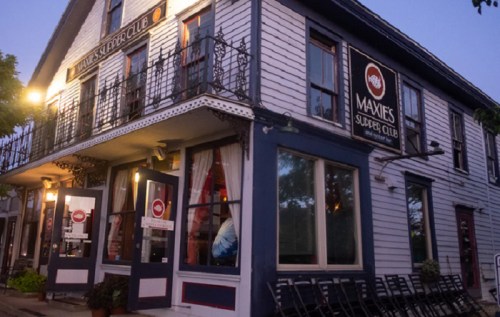 A cozy restaurant exterior at dusk, featuring a sign for "Maxie's Supper Club" and outdoor seating.