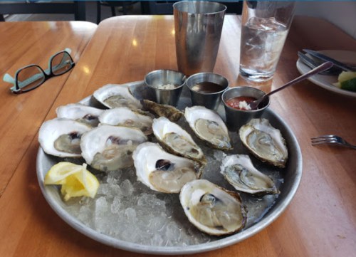 A platter of fresh oysters on ice, accompanied by sauces and a lemon wedge, on a wooden table.