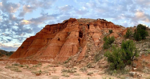 A rugged red rock formation under a cloudy sky, surrounded by sparse vegetation in a desert landscape.