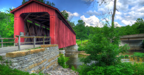 A red covered bridge stands over a calm stream, surrounded by lush greenery and a clear blue sky.
