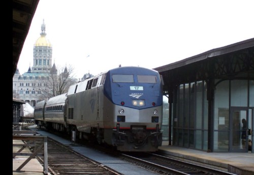 A train arrives at a station with a historic building in the background, featuring a dome and cloudy sky.