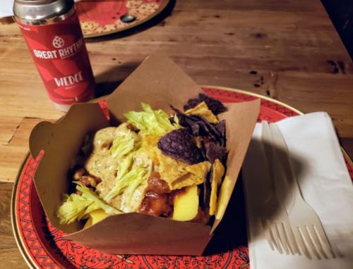 A tray of nachos topped with lettuce, cheese, and bacon, alongside a can of beer and utensils on a wooden table.