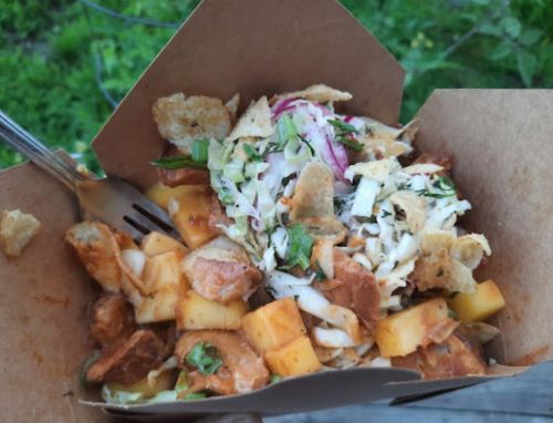 A takeout box filled with diced potatoes, mango, coleslaw, and crispy toppings, set against a green background.