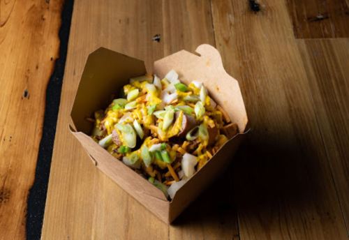 A heart-shaped container filled with nachos topped with cheese, green onions, and diced vegetables on a wooden surface.
