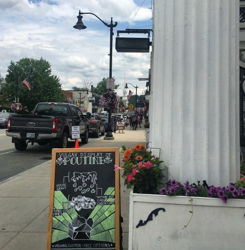 A sidewalk scene featuring a sign for a restaurant with a poutine menu, surrounded by flowers and a busy street.