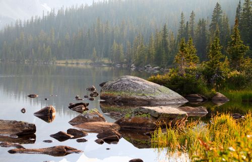 A serene lake surrounded by lush trees and rocky shores, reflecting the calm water and misty mountains in the background.