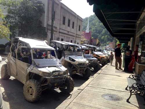 A row of off-road vehicles parked along a sidewalk in a small town, with people nearby and hills in the background.