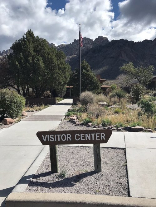 A sign reading "Visitor Center" in front of a building, with mountains and a flag in the background under a cloudy sky.