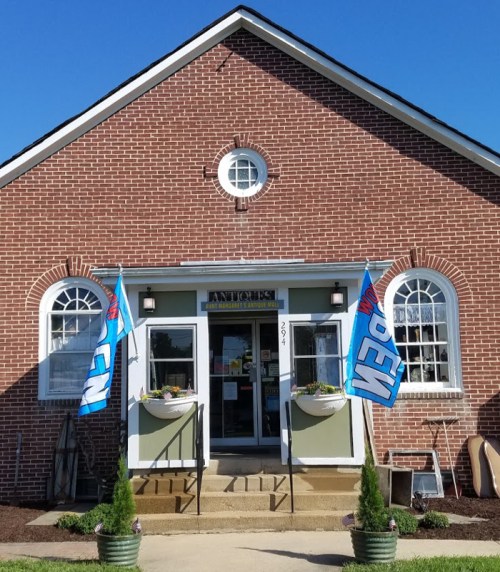 A brick building with a welcoming entrance, featuring blue "OPEN" flags and planters on either side of the door.