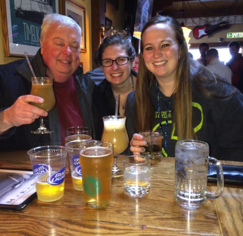 A group of three people smiling at a bar, surrounded by various drinks on the table.