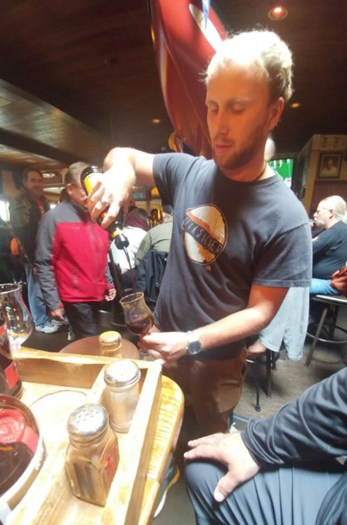 A man in a black shirt pours a drink at a bar, with patrons and a wooden tray of condiments in the background.