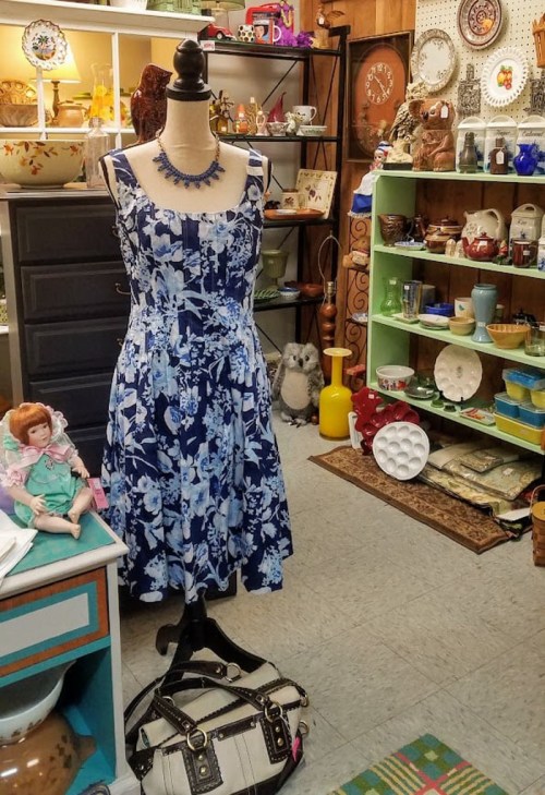 A floral blue dress on a mannequin in a vintage shop, surrounded by various antiques and collectibles.