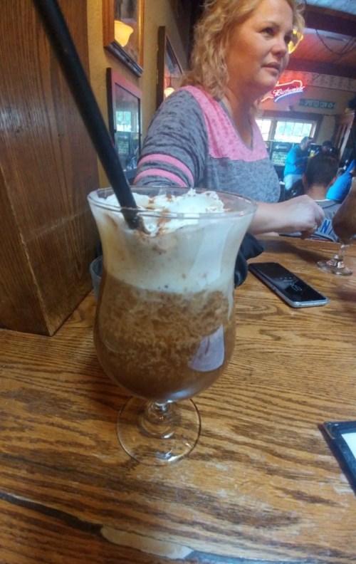 A frosty drink topped with whipped cream sits on a wooden table, with a woman in the background.