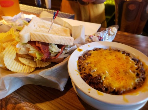 A sandwich with bacon, lettuce, and tomato, served with chips, alongside a bowl of chili topped with melted cheese.