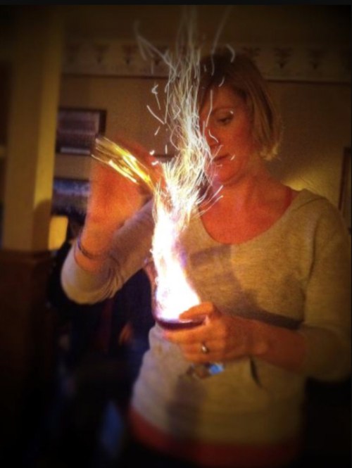 A woman in a gray sweater holds a glass, with bright sparks and flames emerging from it in a dimly lit room.