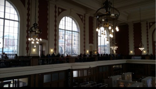 Interior of a large building with tall windows, chandeliers, and a line of people along the balcony.