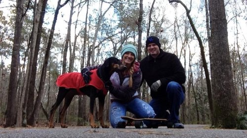 A woman and a man kneel beside a dog in a red coat on a forest path, smiling and enjoying the outdoors.