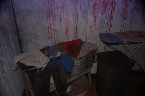 A dimly lit room with a laundry basket filled with clothes, a trash bin, and red streaks on the walls.