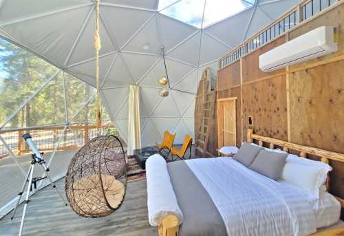 Cozy geodesic dome interior featuring a bed, hanging chair, telescope, and large windows with natural light.