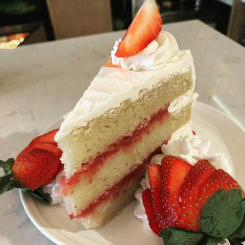 A slice of vanilla cake with strawberry filling, topped with whipped cream and fresh strawberries on a white plate.