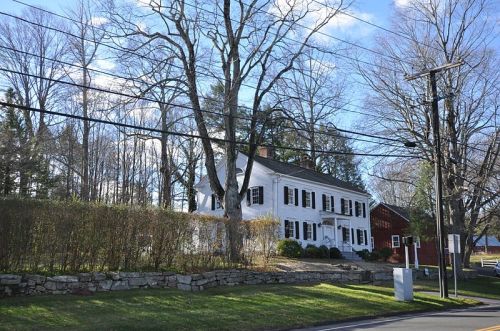 A white colonial-style house with black shutters, surrounded by trees and power lines, set against a clear blue sky.