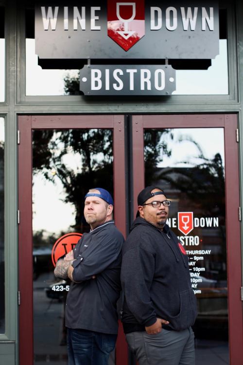 Two men stand back-to-back outside the Wine Down Bistro, with the restaurant's logo visible above them.
