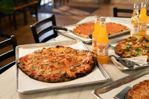 A table with several pizzas on trays and two bottles of orange soda in a casual dining setting.