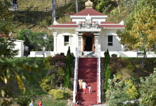A white building with a red roof and stairs, surrounded by greenery and people walking nearby.