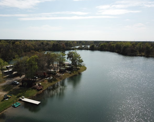 Aerial view of a serene lake surrounded by trees and a small campsite along the shore under a clear sky.