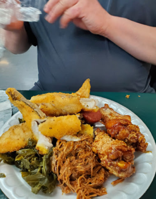 A plate of food featuring fried fish, fried chicken, pulled pork, and collard greens, with a hand reaching for a drink.