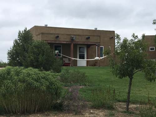 A single-story adobe-style house surrounded by greenery and a grassy area under a cloudy sky.