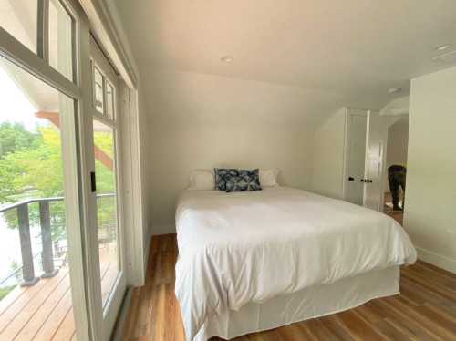 Bright bedroom with a large bed, white linens, and a decorative pillow, featuring windows and wooden flooring.