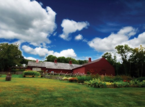 A red barn surrounded by lush greenery and colorful gardens under a blue sky with fluffy clouds.