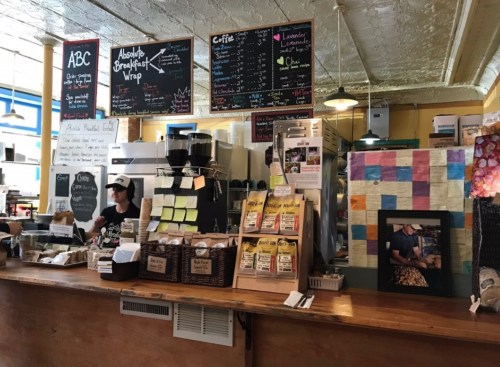 A cozy café interior with a counter, menu boards, and a barista preparing drinks. Baskets of goods are displayed.