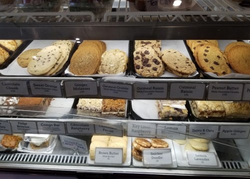 A display case filled with various cookies, including chocolate chunk, oatmeal raisin, and peanut butter, labeled with prices.