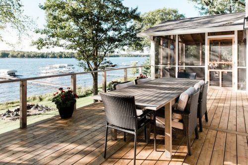 A wooden deck with a dining table and chairs overlooks a serene lake with boats and trees in the background.