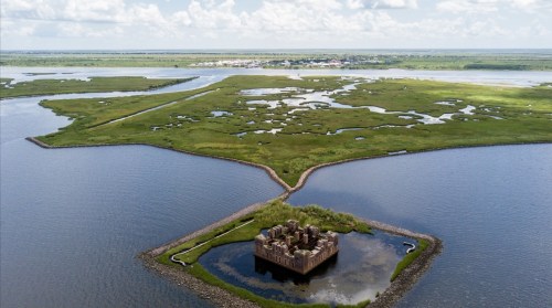 Aerial view of a historic structure surrounded by water and lush green marshlands under a bright sky.