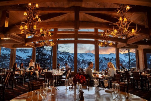 Cozy restaurant with large windows showcasing snowy mountains, warm lighting, and diners enjoying their meals.