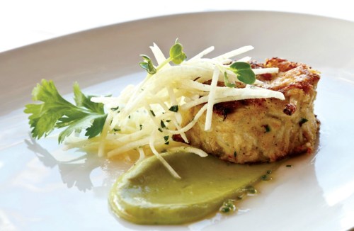 A plated crab cake topped with julienned vegetables and a green sauce, garnished with herbs.