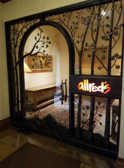 A decorative iron gate with floral designs, leading to a warmly lit room with wooden furniture. The sign reads "allred's."