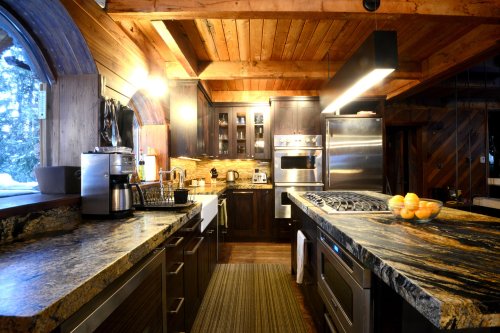 A cozy kitchen with wooden beams, dark cabinets, granite countertops, and modern appliances, featuring a bowl of oranges.