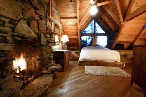 Cozy cabin bedroom with a stone fireplace, wooden walls, and a large window overlooking trees. Warm and inviting atmosphere.