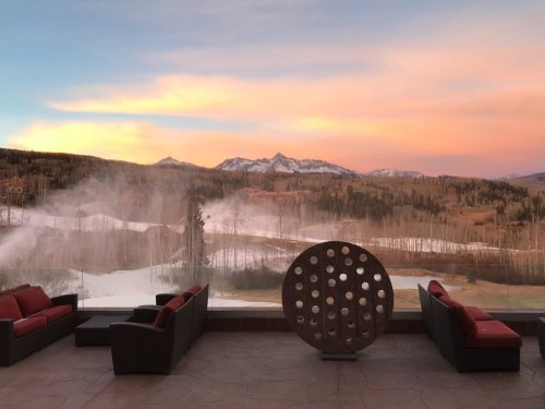 A scenic view of mountains at sunset, with mist rising and modern seating in the foreground.