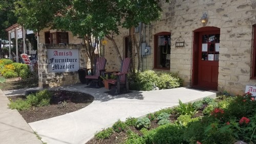 Exterior of Amish Furniture Market with stone walls, a sign, and wooden chairs on a landscaped path.