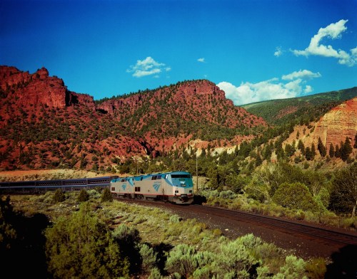A train travels along a scenic route with red rock formations and green trees under a blue sky with fluffy clouds.