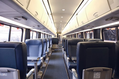 Interior of a train carriage featuring rows of blue seats and overhead storage compartments.