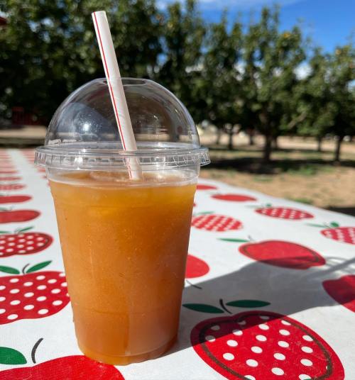 A clear cup filled with orange drink and a straw, set on a table with a red apple pattern, surrounded by trees.