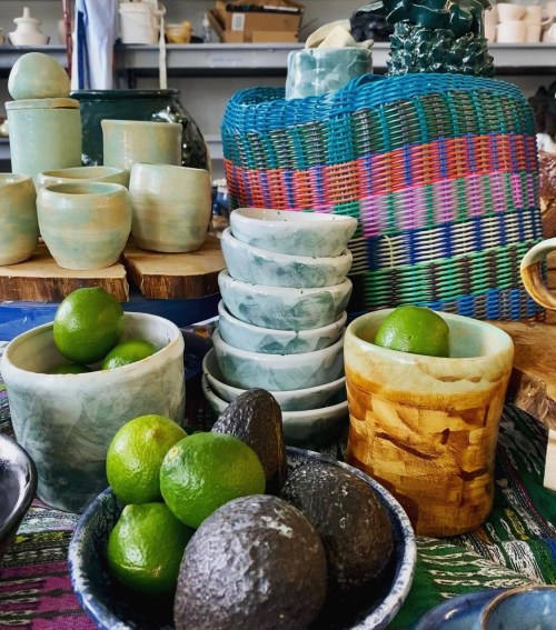 A colorful display of pottery, including cups and bowls, with limes and avocados arranged on a vibrant tablecloth.